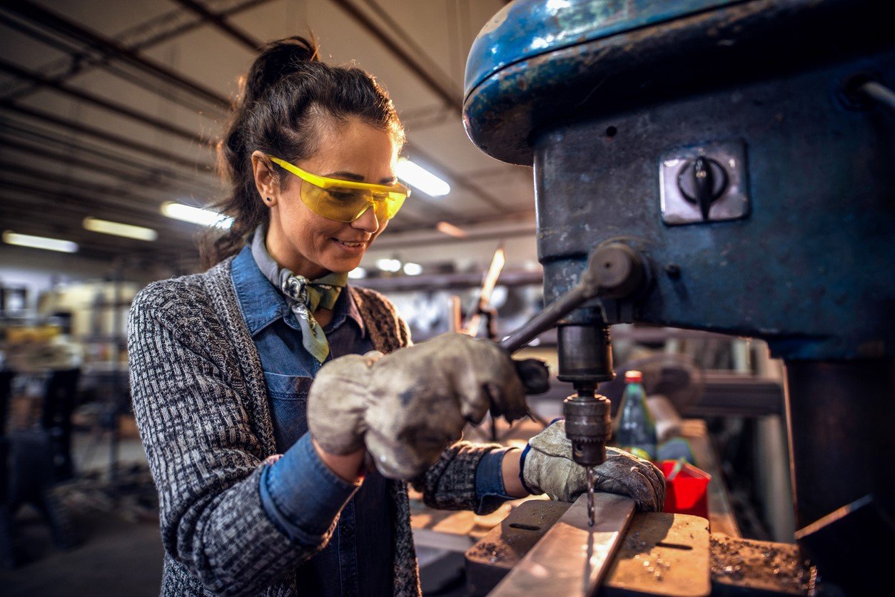 Vrouw met veligheidsbril staat aan machine die een gat boort in een metalen lat